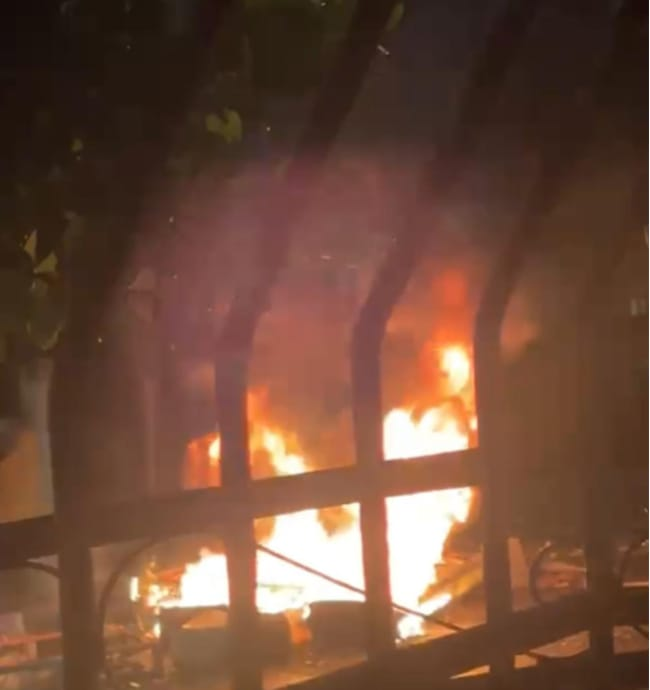Llamaradas intensas provocadas por la quema de neumáticos en la avenida Independencia de Pimentel durante la noche, captadas a través de las rejas de hierro de una vivienda.
