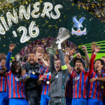 El delantero Jean-Philippe Mateta junto a sus compañeros del Crystal Palace levantando el trofeo de campeones de la UEFA Conference League en el podio de Leipzig, rodeados de confeti y celebrando de manera eufórica.